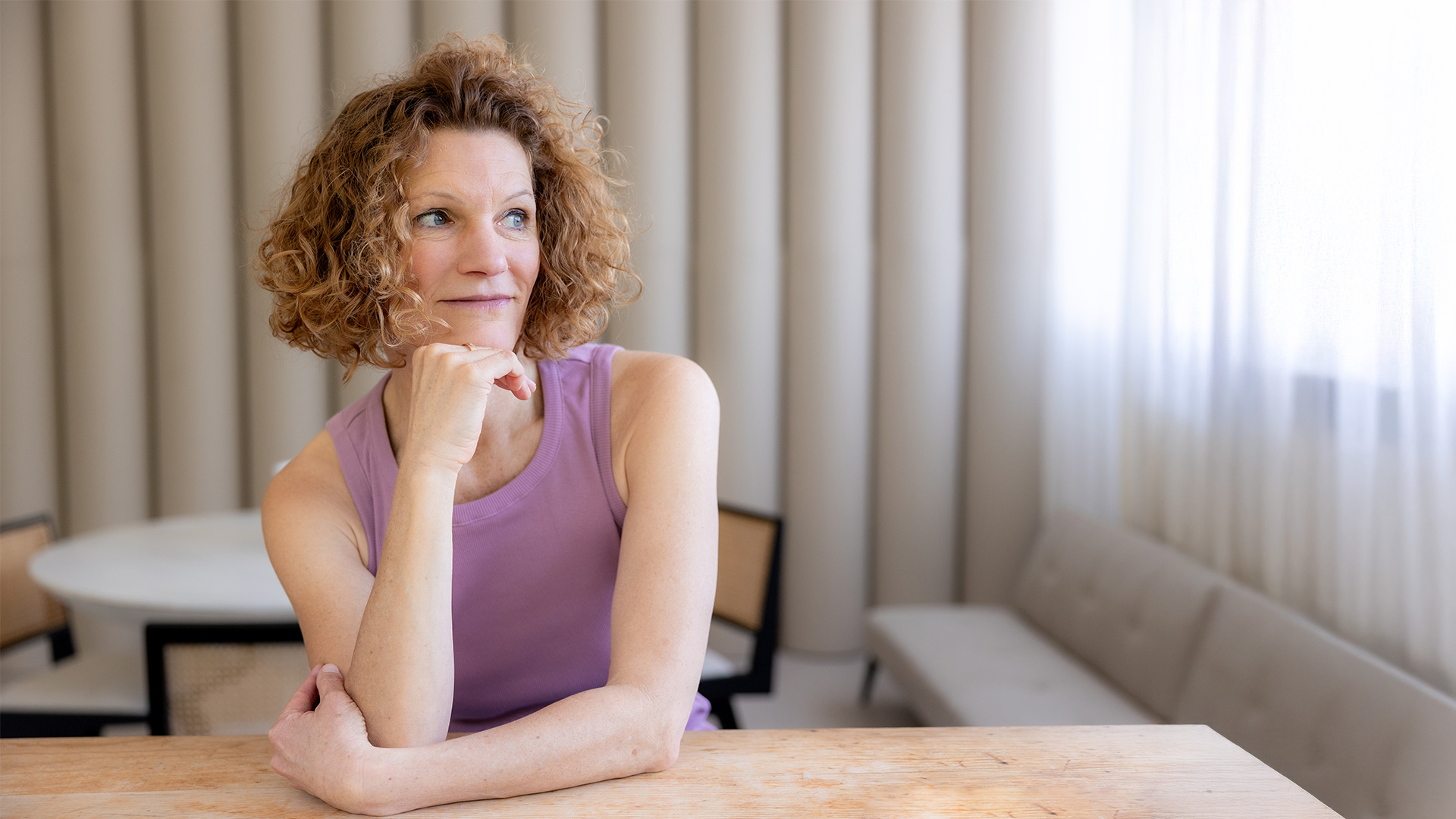 Frau sitzt an Holztisch, Kinn auf rechte Hand aufgestützt, Arme liegen auf dem Tisch, schaut in Richtung Fenster